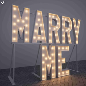 Op de afbeelding: Grote, verlichte letters die "MARRY ME" spellen. De witte letters zijn omlijnd met warme, gloeiende gloeilampen. Het bord is gemonteerd op een wit metalen frame, geschikt voor een aanzoek of bruiloft. De letters zijn ongeveer 100 cm hoog.