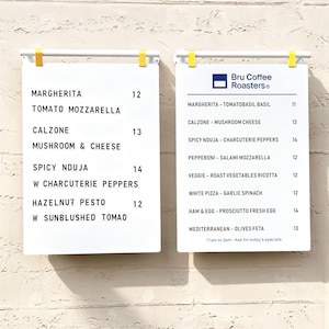 May include: Two white rectangular menu boards with black lettering suspended by white bars and yellow clips. The left board lists pizza options like Margherita and Calzone. The right board features the Bru Coffee Roasters logo and menu items.