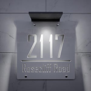 以下が含まれることがあります： モダンな照明付き住所サイン。このサインは、数字「2117」が切り抜かれ、その下に通り名「Rosecliff Road」が記された、明るい灰色の長方形です。大理石のような壁に取り付けられ、上部に黒い照明器具が付いています。