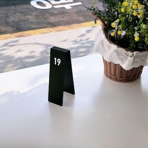 May include: A black A-frame table number sign with the white number 19 printed on the front. The sign is made of a smooth, matte material and stands upright on a flat surface, positioned next to a brown woven basket filled with flowers.