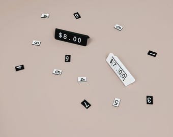 May include: Two angled price display stands, one black with white text reading $8.00 and one white with black text reading $7.00, surrounded by scattered individual rectangular tiles featuring single black or white digits.