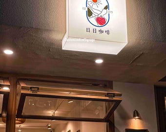 May include: A white illuminated sign with the text "Daily Coffee" in a circle with a sun and mountains inside. The text "Est. 2019" is below the circle. The text "每日 咖啡" is below the circle in Japanese. The sign is mounted on a brown ceiling.