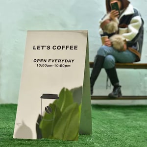 Może przedstawiać: Dwustronny szyld z napisem "LET'S COFFEE" i "OTWARTE CODZIENNIE 10:00-22:00". Szyld przedstawia grafikę filiżanki kawy. Szyld jest biały z zielonym tłem. W tle osoba siedząca na ławce.