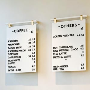 Peut inclure: Deux panneaux rectangulaires blancs avec du texte noir énumérant les options de café et d'autres boissons. Les panneaux sont suspendus par du matériel doré. Le menu du café comprend l'espresso, l'americano et le cappuccino. L'autre menu comprend le lait doré, le chocolat chaud et le matcha latte.