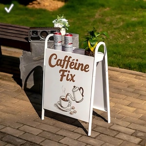 Könnte beinhalten: Ein weißes A-förmiges Schild mit den Worten "Caffeine Fix" in braunen Buchstaben. Das Schild zeigt eine Illustration einer Kaffeetasse und einer Teekanne. Zwei Bierdosen und eine kleine Pflanze stehen auf dem Schild. Ein Lautsprecher und eine Bank befinden sich im Hintergrund.