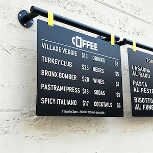 May include: A black rectangular menu board hanging from a black metal rod with yellow clips. The board features white text listing items like Village Veggie, Turkey Club, Bronx Bomber, Pastrami Press, Spicy Italiano, and various drinks with prices.