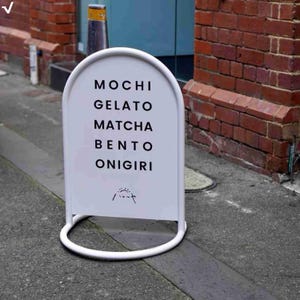 Könnte beinhalten: Ein weißes, bogenförmiges Werbeschild mit schwarzem Text, der "MOCHI, GELATO, MATCHA, BENTO, ONIGIRI" auflistet. Das Schild hat einen weißen Metallrahmen und -fuß. Der Hintergrund umfasst eine Backsteinmauer und einen Teil einer blauen Tür.