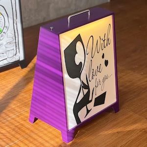 May include: A purple A-frame sign with a silver handle. The sign has a white illuminated panel with black graphic art and the words "With love for you...". The sign is on a wooden surface.