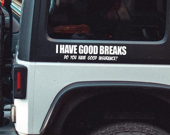 May include: A white Jeep SUV with black tires and rims is parked on a city street. A spare tire is mounted on the back. A white decal on the rear window reads, "I HAVE GOOD BREAKS. DO YOU HAVE GOOD INSURANCE?"