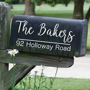 Peut inclure: Boîte aux lettres noire avec inscription blanche "The Bakers" et "92 Holloway Road". La boîte aux lettres est montée sur un poteau en bois et entourée de fleurs blanches.