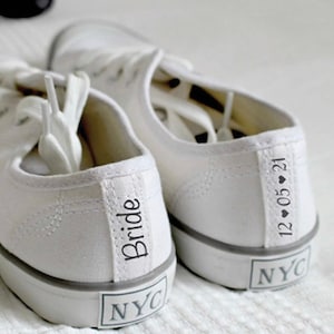 May include: A pair of white canvas sneakers with the word "Bride" and the date "12.05.21" written on them. The sneakers also have the letters "NYC" written on them.