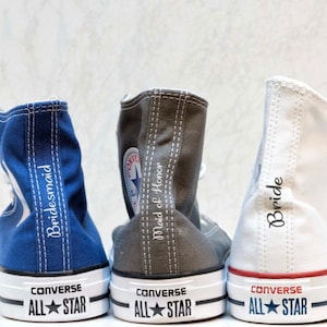 Puede incluir: Tres pares de zapatillas Converse de caña alta con bordado personalizado. Las zapatillas son azules, grises y blancas. Las zapatillas azules dicen "Bridemaid", las grises dicen "Maid of Honor" y las blancas dicen "Bride".