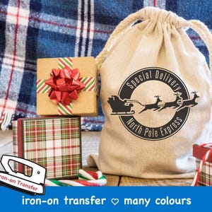 Pode incluir: Design de transferência de ferro com um Papai Noel em um trenó puxado por renas com o texto "Special Delivery North Pole Express". O design está em uma bolsa de lona branca com caixas de presente xadrez vermelhas e verdes e bastões de doces.