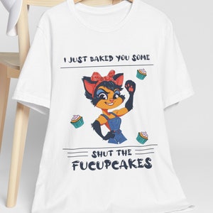 May include: White t-shirt with a cartoon cat wearing a red bandana and blue overalls. The cat is holding up a fist and there are cupcakes around her. The text on the shirt reads "I just baked you some SHUT THE FUCUPCAKES".