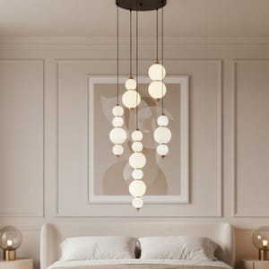 May include: A modern bedroom featuring a cream-colored bed with a neutral-toned comforter and pillows. Above the bed hangs a stylish chandelier with multiple spherical, frosted glass lights. Two matching bedside tables with lamps complete the elegant decor.