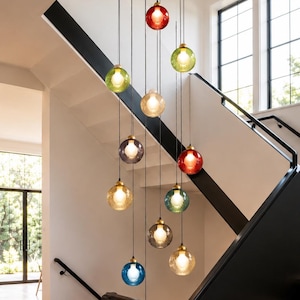 May include: A multi-light pendant chandelier featuring twelve spherical glass globes in various colours including blue, red, green, yellow, and clear. Each globe is suspended by a thin black cord from a central fixture, creating a cascading arrangement.