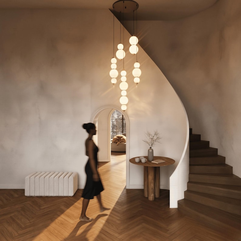 May include: A multi-pendant chandelier featuring several vertical arrangements of glowing white spherical glass globes suspended from a circular ceiling mount, casting a warm light over a wooden floor and a curved staircase.