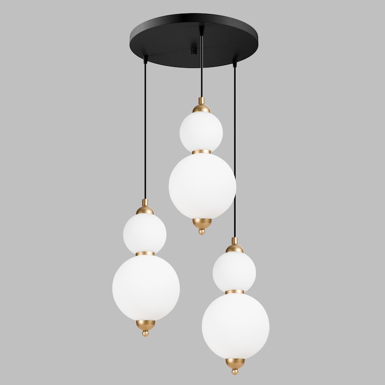 May include: A modern pendant light fixture with three white globe shades suspended from a black circular canopy. The shades are accented with gold-colored hardware. The light fixture is designed for indoor use and adds a touch of elegance to any room.