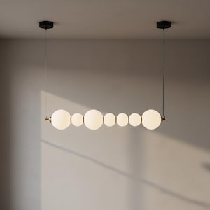 May include: A modern, linear chandelier featuring a series of white, spherical glass shades of varying sizes. The fixture is suspended from the ceiling by thin wires and has black ceiling mounts. The metal accents are gold-toned.