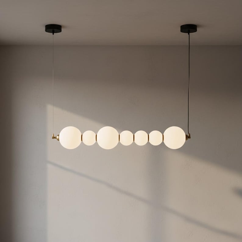 May include: A modern, linear chandelier featuring a series of white, spherical glass shades of varying sizes. The fixture is suspended from the ceiling by thin wires and has black ceiling mounts. The metal accents are gold-toned.