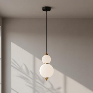 May include: A modern pendant light fixture with two frosted glass globes stacked on top of each other. The fixture has brass-colored accents and a black ceiling mount. The light hangs from a black cord.