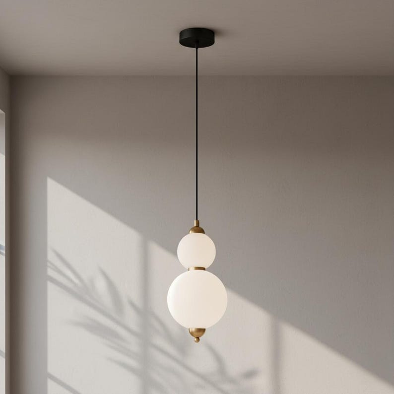 May include: A modern pendant light fixture with two frosted glass globes stacked on top of each other. The fixture has brass-colored accents and a black ceiling mount. The light hangs from a black cord.