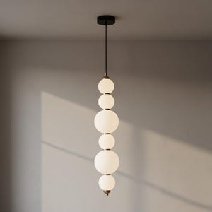 May include: A modern pendant light fixture with a black ceiling mount and a series of white, spherical glass shades. The shades are connected by brass-colored metal accents, creating a vertical, elongated design. The light hangs from a black cord.