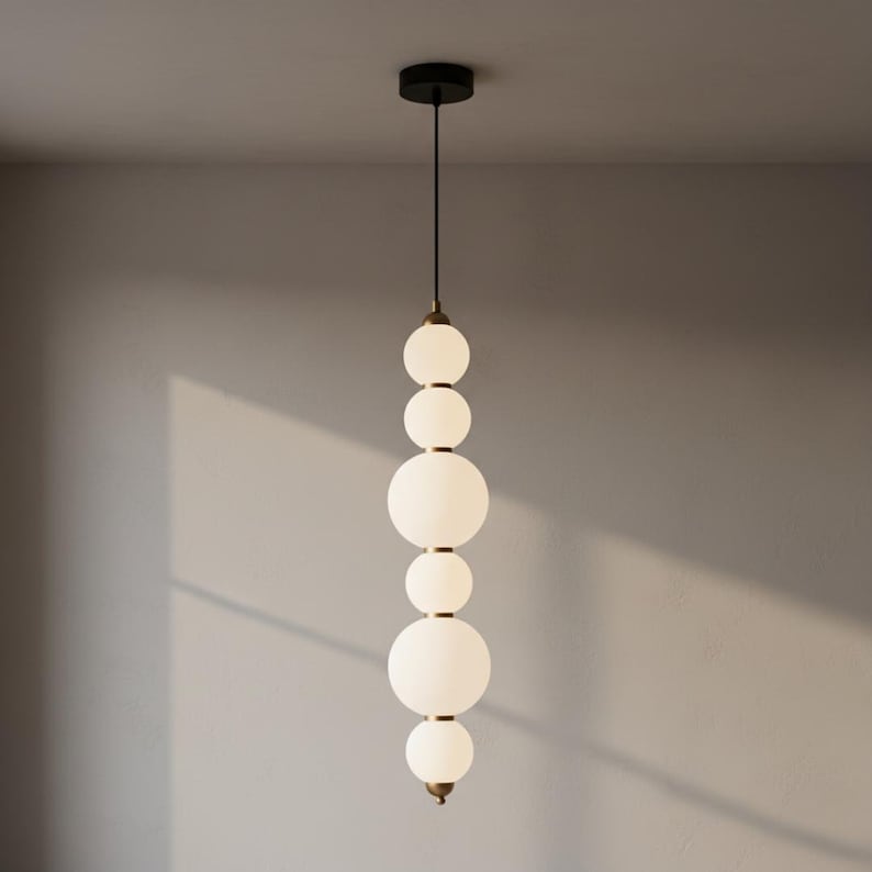 May include: A modern pendant light fixture with a black ceiling mount and a series of white, spherical glass shades. The shades are connected by brass-colored metal accents, creating a vertical, elongated design. The light hangs from a black cord.