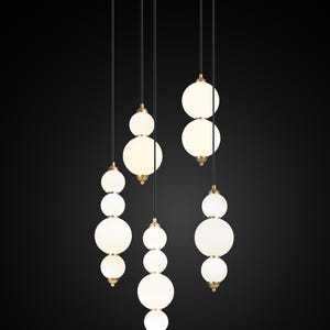 May include: Modern pendant light fixture with multiple white globe shades of varying sizes. The lights hang from a circular, dark gray ceiling plate. Each globe is suspended by a black cord and accented with a gold-colored metal detail.