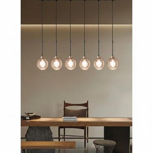 Puede incluir: Un comedor moderno con una larga mesa de madera y seis lámparas colgantes. Cada lámpara presenta una pantalla de vidrio texturizado en forma de globo con un cálido tono ámbar. Las luces cuelgan de cables negros, creando un ambiente elegante.
