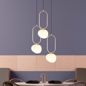 May include: Three white glass globe pendant lights with gold metal frames hanging from the ceiling. The lights are hanging over a table with chairs in a dining room.