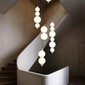 Modern staircase chandelier with staggered hanging white opal glass globes from a round metal ceiling plate, softly glowing over a tall stairwell with warm ambient light.