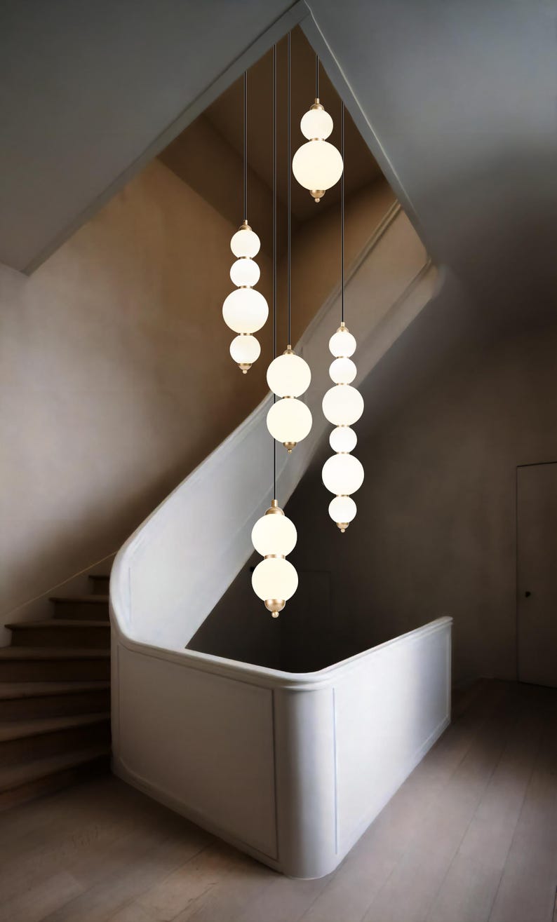 Modern staircase chandelier with staggered hanging white opal glass globes from a round metal ceiling plate, softly glowing over a tall stairwell with warm ambient light.