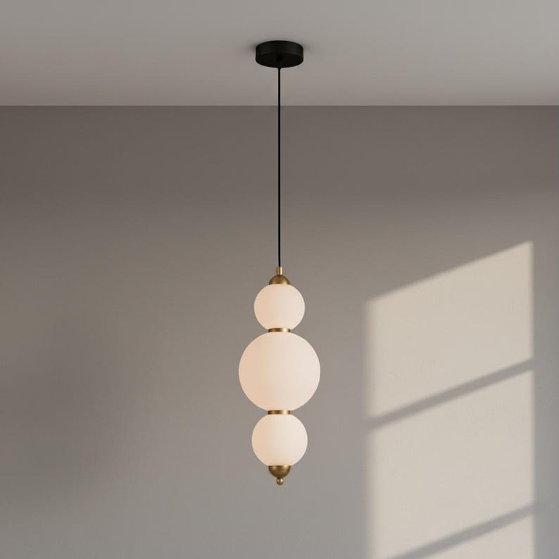 May include: A modern pendant light fixture with three frosted glass globes of varying sizes. The globes are stacked vertically and connected by brass-colored accents. The fixture hangs from a black ceiling mount and a black cord.