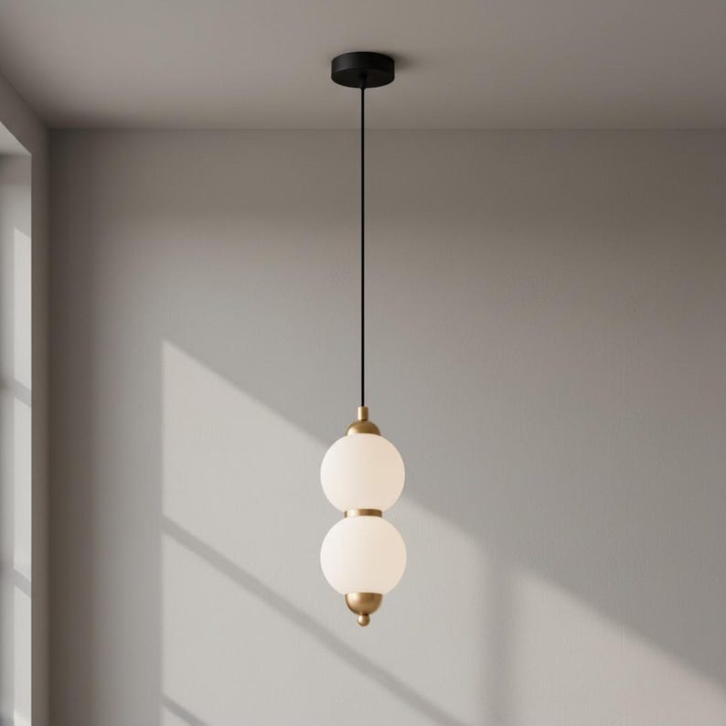May include: A modern pendant light with two spherical, frosted glass shades stacked atop each other. The fixture has brass-colored accents and a black ceiling mount. The light hangs from a black cord, creating a minimalist design.