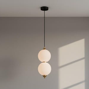 May include: A modern pendant light with two frosted glass globes stacked vertically. The fixture has a black ceiling mount, a black cord, and brass-colored accents. The light is suspended against a neutral gray wall.