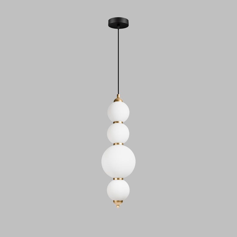 May include: A modern pendant light with a black ceiling mount and a black cord. The light fixture features four white, spherical glass shades of varying sizes, accented with gold-colored metal rings and end caps. The design is minimalist and elegant.
