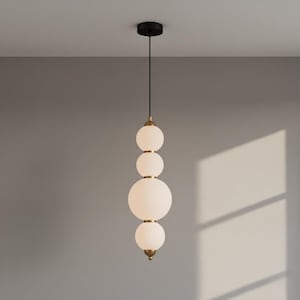 May include: A modern pendant light fixture with four frosted glass globes of varying sizes, suspended from a black ceiling mount. The globes are connected by brass-colored rings and a brass-colored finial at the bottom. The light fixture hangs from a black cord.