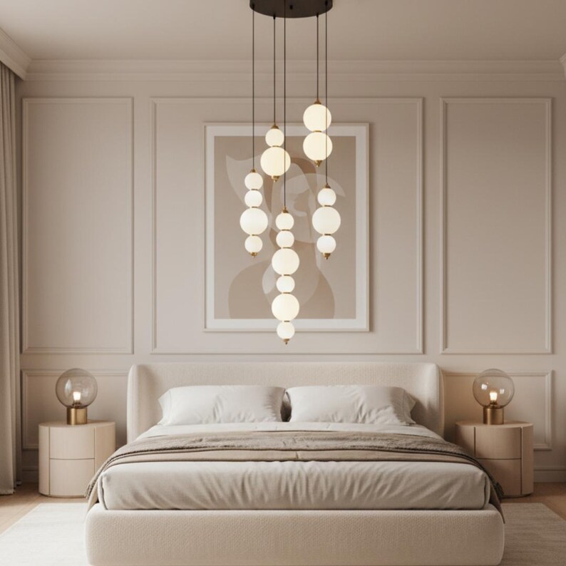 May include: A modern bedroom featuring a cream-colored bed with a neutral-toned comforter and pillows. Above the bed hangs a stylish chandelier with multiple spherical, frosted glass lights. Two matching bedside tables with lamps complete the elegant decor.