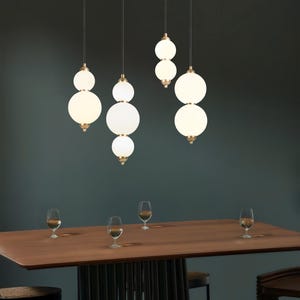 May include: Several modern pendant lights with white globe shades hang above a wooden dining table. The lights have a brass finish and are suspended from black cords. Wine glasses are arranged on the table.