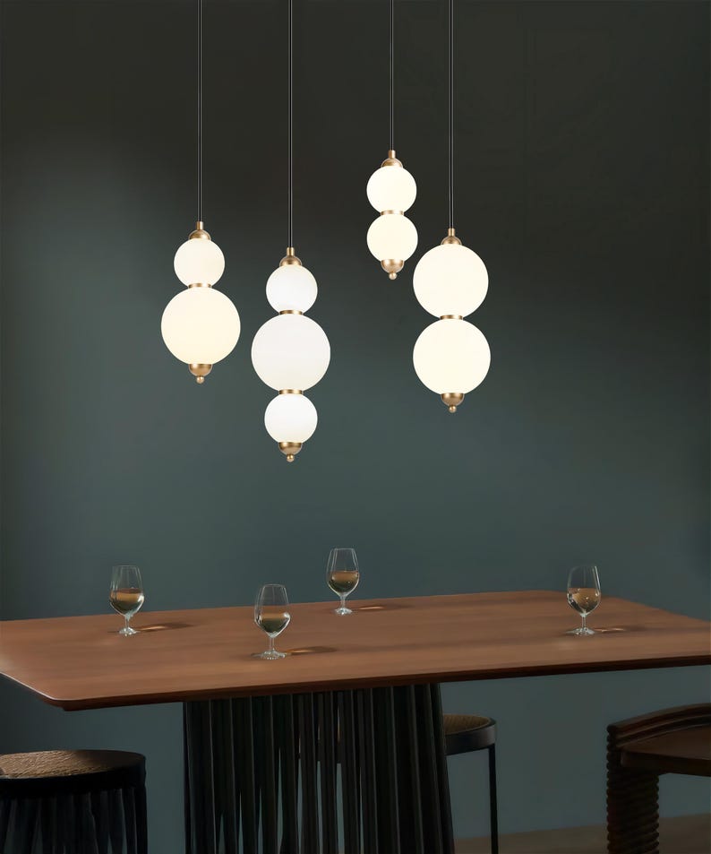 May include: Several modern pendant lights with white globe shades hang above a wooden dining table. The lights have a brass finish and are suspended from black cords. Wine glasses are arranged on the table.