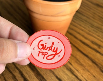 May include: An oval-shaped sticker with a light pink background and a red border. The sticker features the red handwritten text "Girly Pop" in the center, surrounded by four small red "x" marks placed symmetrically around the lettering.