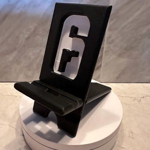 May include: A black plastic phone stand with a cut-out of the Rainbow Six Siege logo. The stand is angled to hold a phone upright.