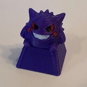 May include: A purple 3D printed mechanical keyboard keycap featuring a stylized creature with a round body, pointed ears, red eyes, and a wide white grin. The figure sits atop a square purple base, displaying visible horizontal layer lines.