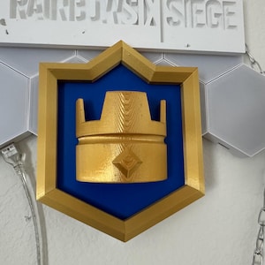 May include: A gold-colored crown emblem on a blue background, set within a gold-edged shield. The emblem is part of a larger wall decoration, with the text "RAINBOW SIX SIEGE" visible above. Hexagonal panels and chains are also present.