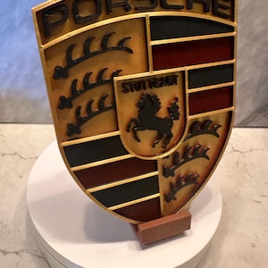 May include: A shield-shaped emblem with the word "PORSCHE" in black lettering at the top. The emblem features gold, black, and red sections, with a smaller shield in the center displaying a black horse. The word "STUTTGART" is also visible.