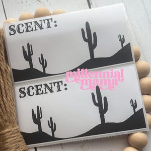 May include: Two white rectangular labels with black cactus silhouettes and the word "SCENT:" printed on them. The bottom label has the words "millennial mama" in pink. The labels are surrounded by wooden beads and twine.