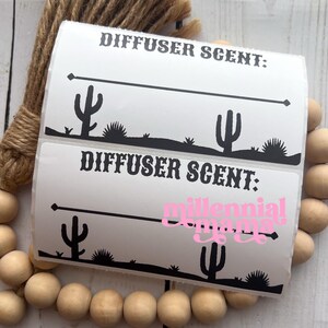 May include: White labels with black cactus and desert landscape designs. The labels read "DIFFUSER SCENT:" and "millennial mama" in pink script. A bundle of brown rope and wooden beads are also in the image.