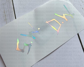May include: A rectangular adhesive decal featuring the letters S.W.D.W. in a stylized, angular font. The lettering has a holographic finish that reflects various colors, including blue, green, and pink, set against a clear, semi-transparent backing.