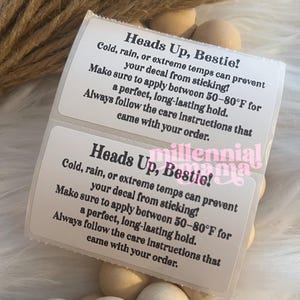 May include: Two white rectangular labels with black text, reading "Heads Up, Bestie!" and instructions for decal application. The labels are on a bed of wooden beads and a fluffy white surface, with a bundle of twine in the background.